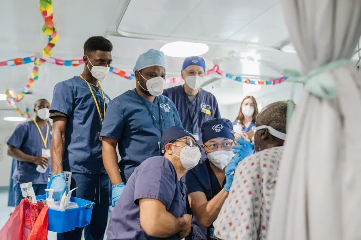 A team of healthcare professionals wearing masks and scrubs attend to a patient in a medical setting, with festive decorations in the background, aboard a Mercy Ship.