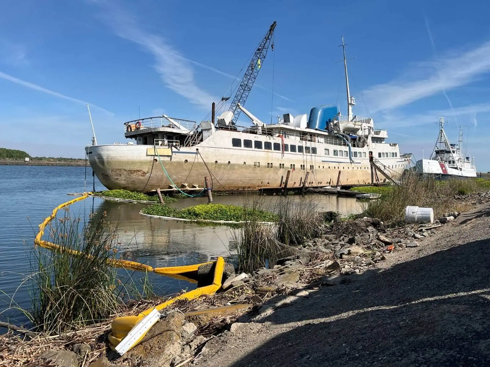 An old ship partially submerged near the shore is surrounded by floating containment booms. A crane and vegetation are visible in the background.