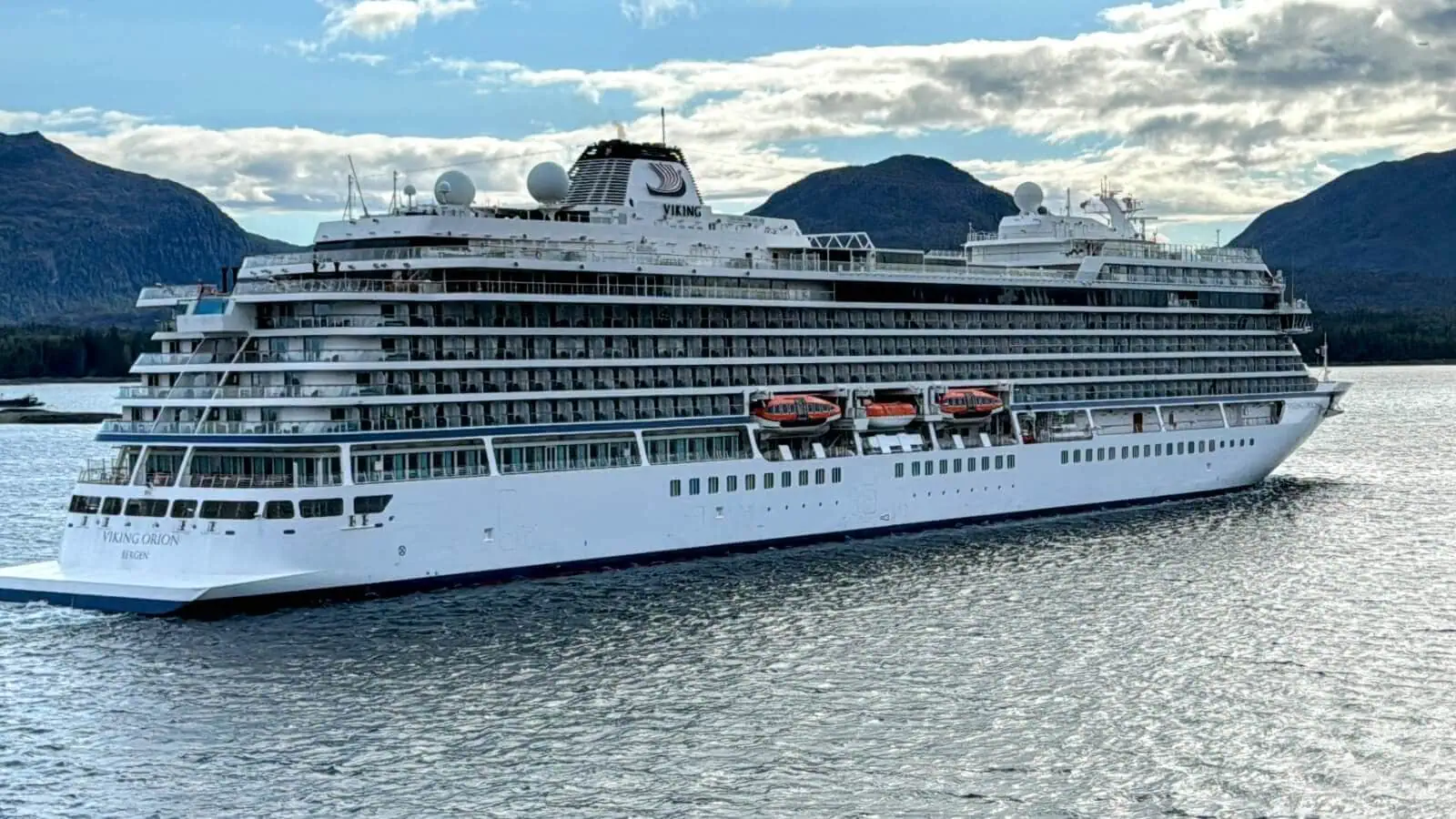 A large white Viking cruise ship sails on calm waters, with green hills and a cloudy sky in the background, showcasing the impressive size of these vessels from smallest to largest.