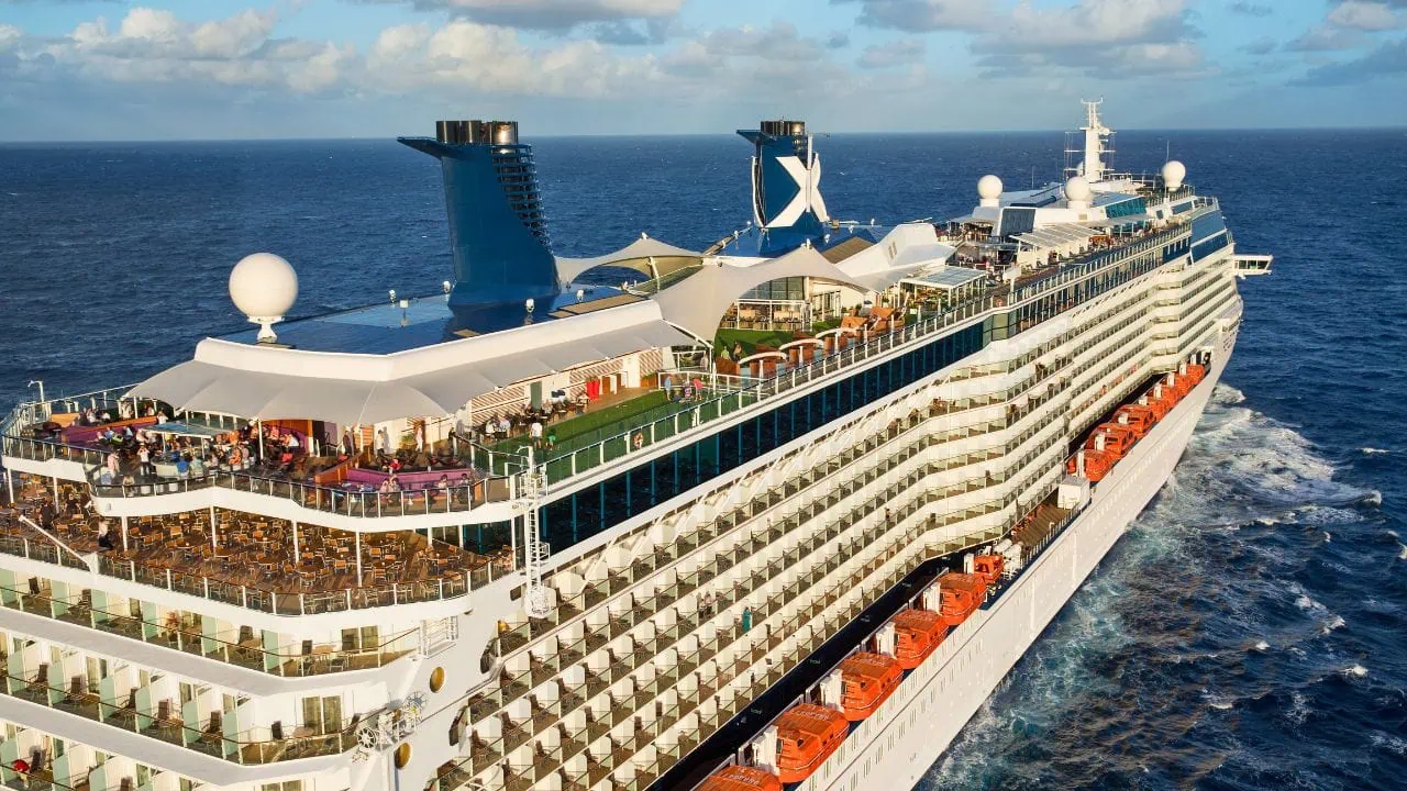 A large cruise ship sailing on the ocean with multiple decks, lifeboats, and a crowd of people on the top deck under blue skies. Enjoy travel savings and cheaper itineraries for 2024 as you embark on this unforgettable journey.