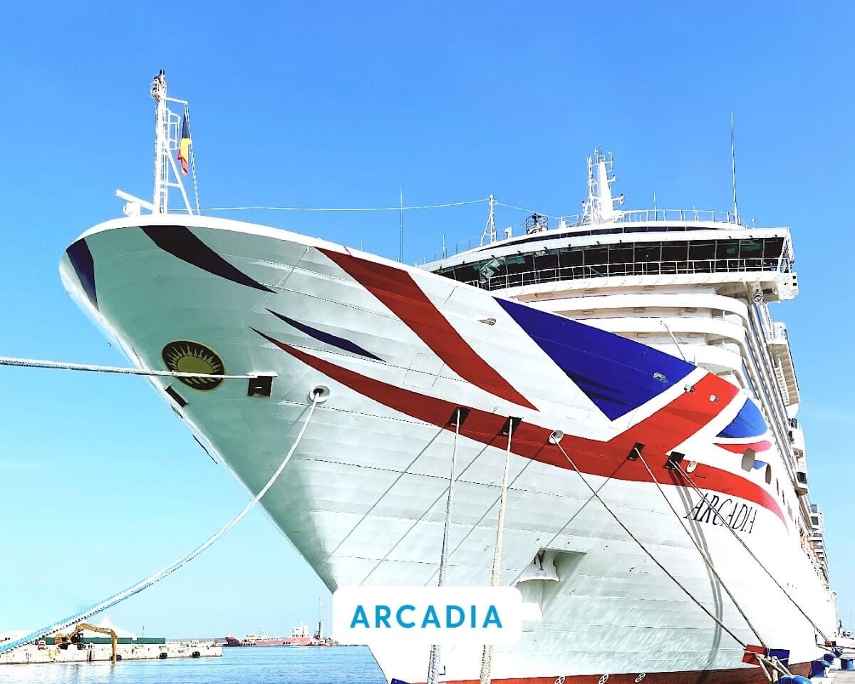 Close-up of the large bow of Arcadia, a P&O Cruise Ship, showcasing its distinctive red, white, and blue pattern. The ship is docked at a UK port under a clear blue sky.