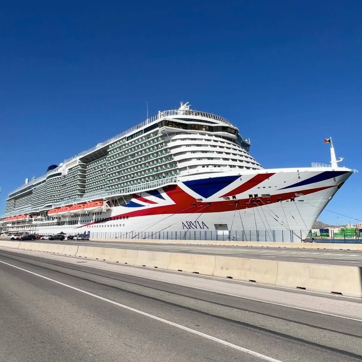 A large P&O Cruise Ship, Arvia, docked at a pier under a clear sky. The ship features a British flag design on its bow and boasts multiple decks with numerous windows and balconies.