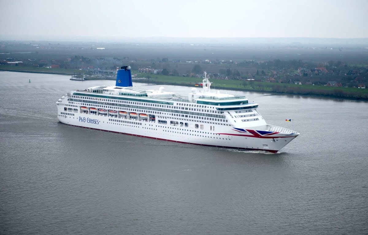 A large white P&O cruise ship from the UK sails on a wide river with a vast landscape and scattered buildings in the background under a cloudy sky.