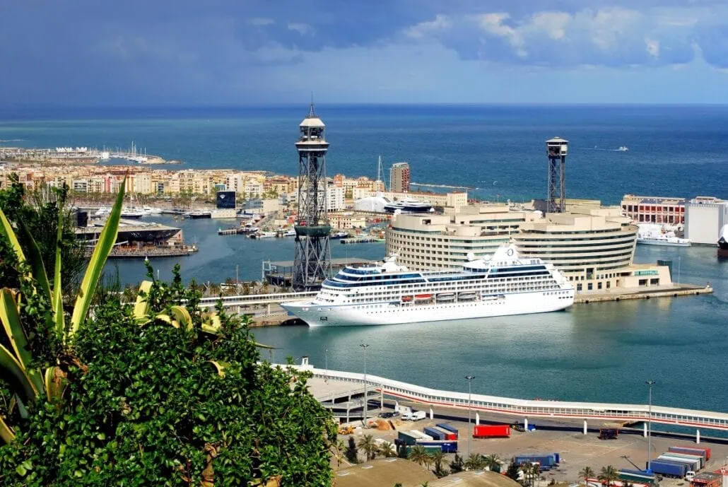 A cruise ship is docked at a European port with industrial buildings and cranes in the background. The harbor is bordered by a city and the sea, with some green plants in the foreground. Despite recent anti-tourist protests, cruise lines continue to bring visitors to this bustling harbor.