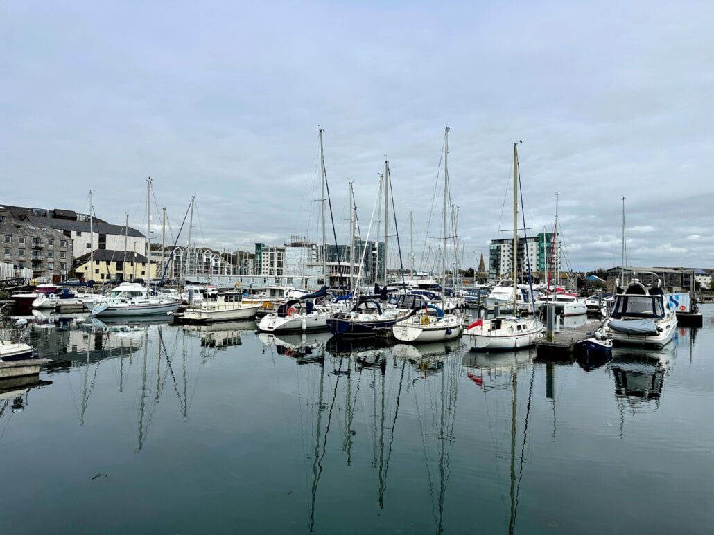 A marina with numerous docked sailboats and yachts on a calm, reflective body of water. The background includes buildings and a cloudy sky, offering picturesque views where you can rent a boat at an affordable price.