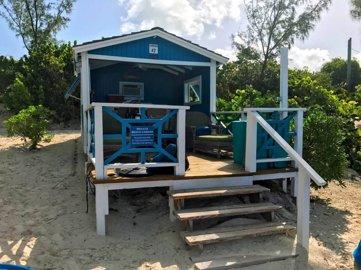 A charming Half Moon Cay cabana, small and blue and white on stilts, features a porch with three steps leading up. Surrounded by lush greenery and soft sand, it sits peacefully under a partly cloudy sky.