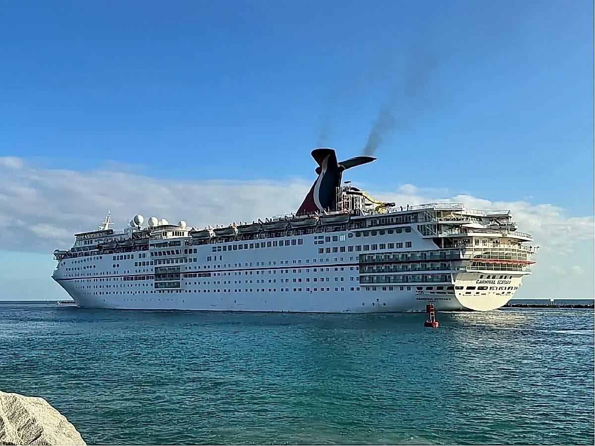 A large white cruise ship with a black and red smokestack, reminiscent of the carnival ships sold years ago, sails through a calm sea under a partly cloudy sky.