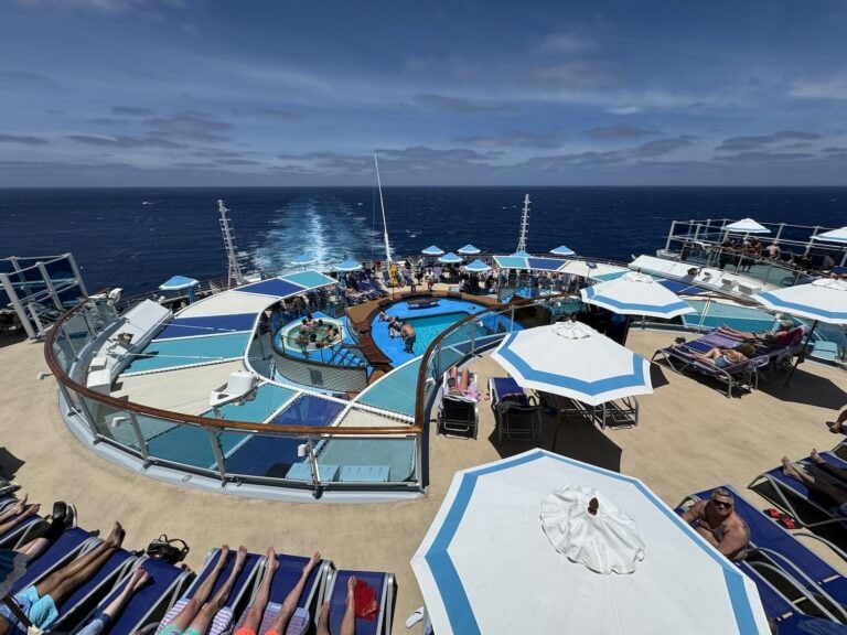 A cruise ship deck with a swimming pool, sunbathers on loungers, and open sea in the background under a clear blue sky.