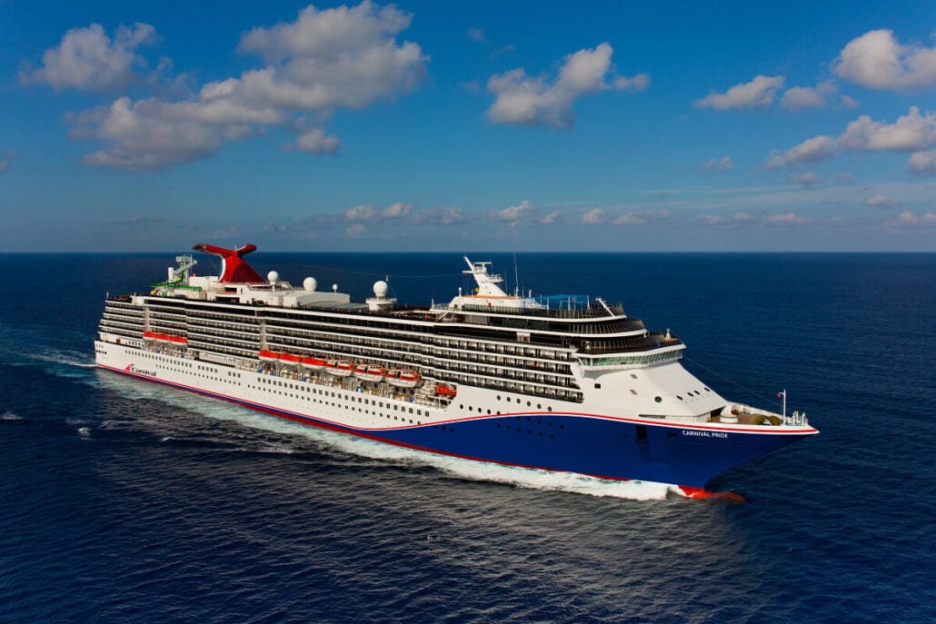 A large cruise ship, the Carnival Pride, with multiple decks sails on the ocean under a partially cloudy sky, en route to the Bahamas.