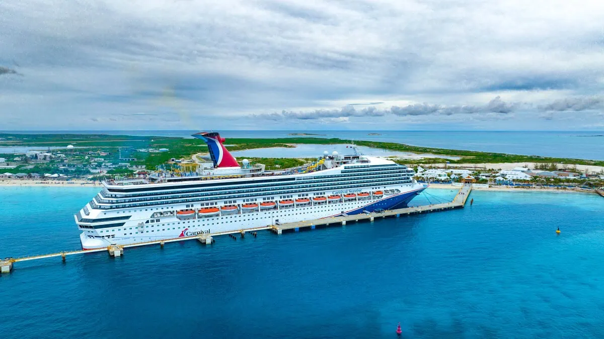 A large cruise ship docked at a pier on a clear blue ocean, with an island in the background under a cloudy sky.