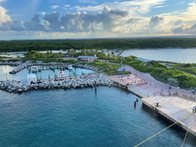 Aerial view of a small marina and docks surrounded by rocky edges and green vegetation on Castaway Cay, with boats docked and a building nearby, under a partly cloudy sky.