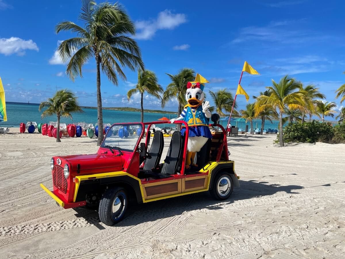 A person in a Donald Duck costume stands in a red and yellow beach vehicle, parked on the sandy shores of Castaway Cay, with palm trees and the ocean in the background.