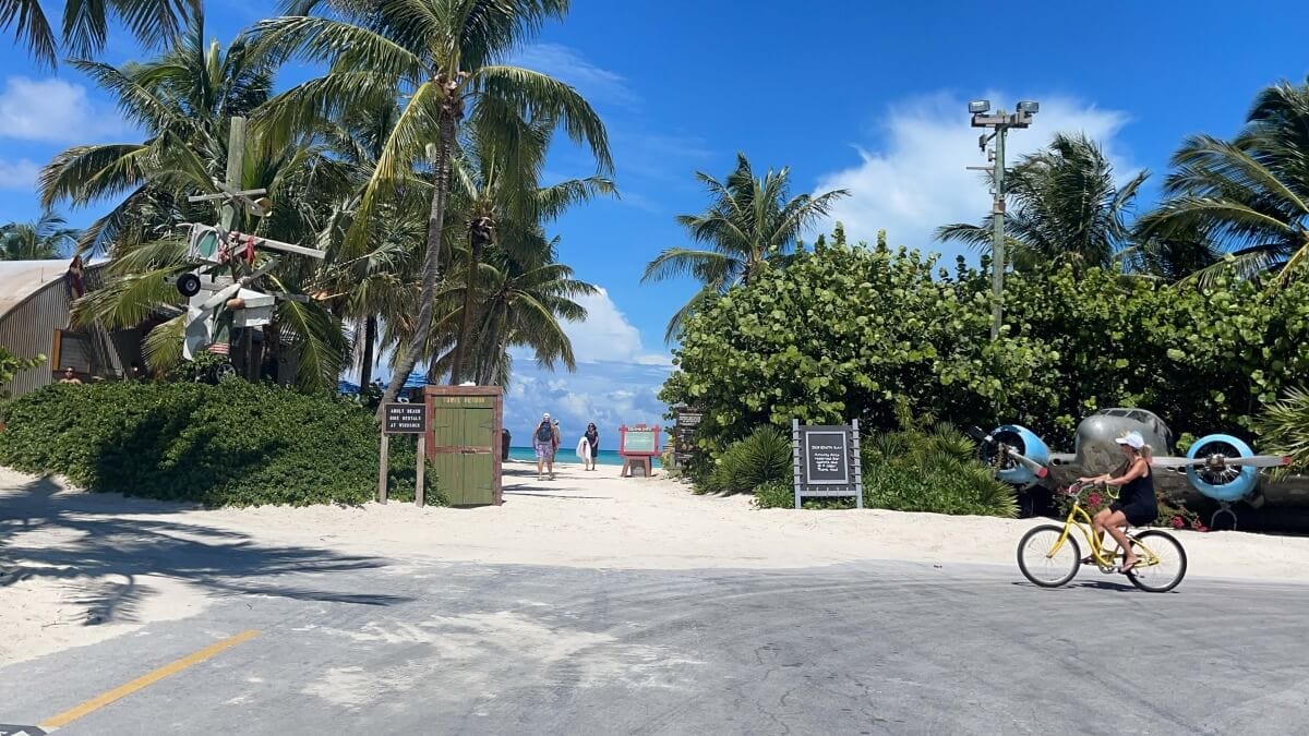 A tropical beach entrance with palm trees, people walking toward the beach, and a person riding a bicycle on a sandy path with vintage airplanes on display evokes the serene charm of Castaway Cay.