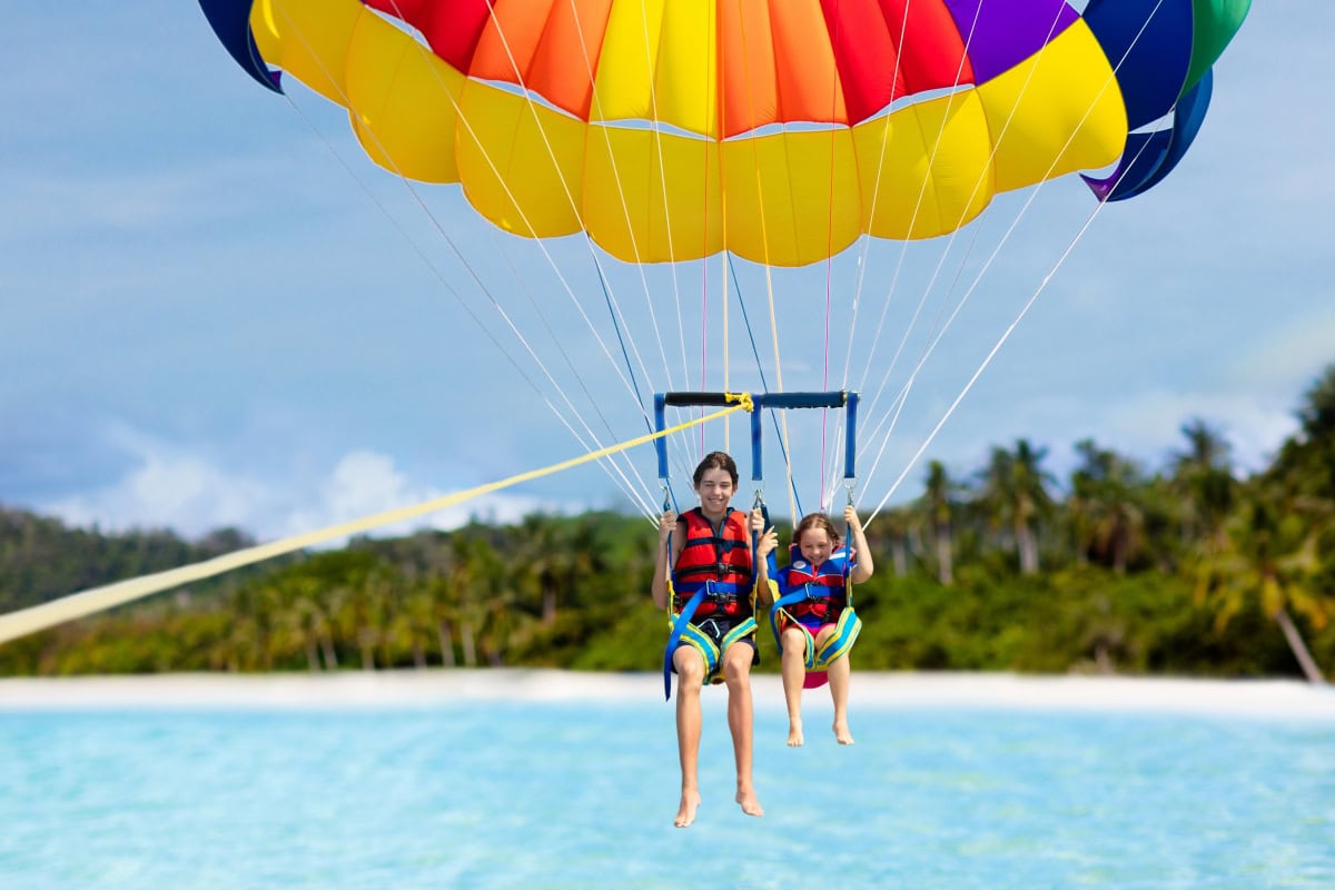 Two people, wearing life jackets, parasailing over the ocean with a colorful parachute. A tropical beach and palm trees are visible in the background, reminiscent of the serene beauty found at Castaway Cay.