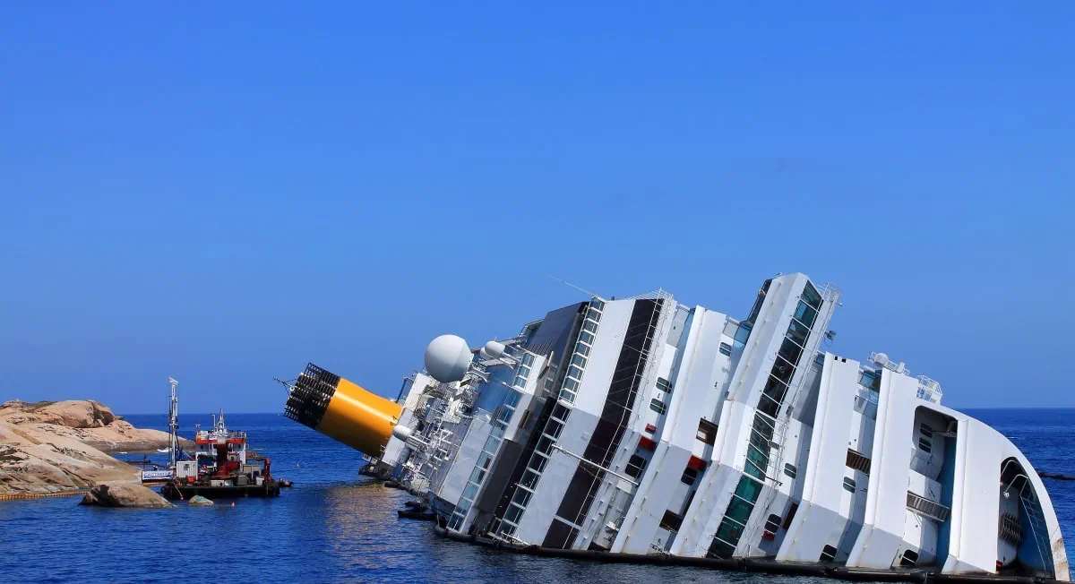 A large white cruise ship is partially submerged and tilting on its side near rocky outcrops in the ocean, raising the question, can a cruise ship tip over? A small boat is nearby.