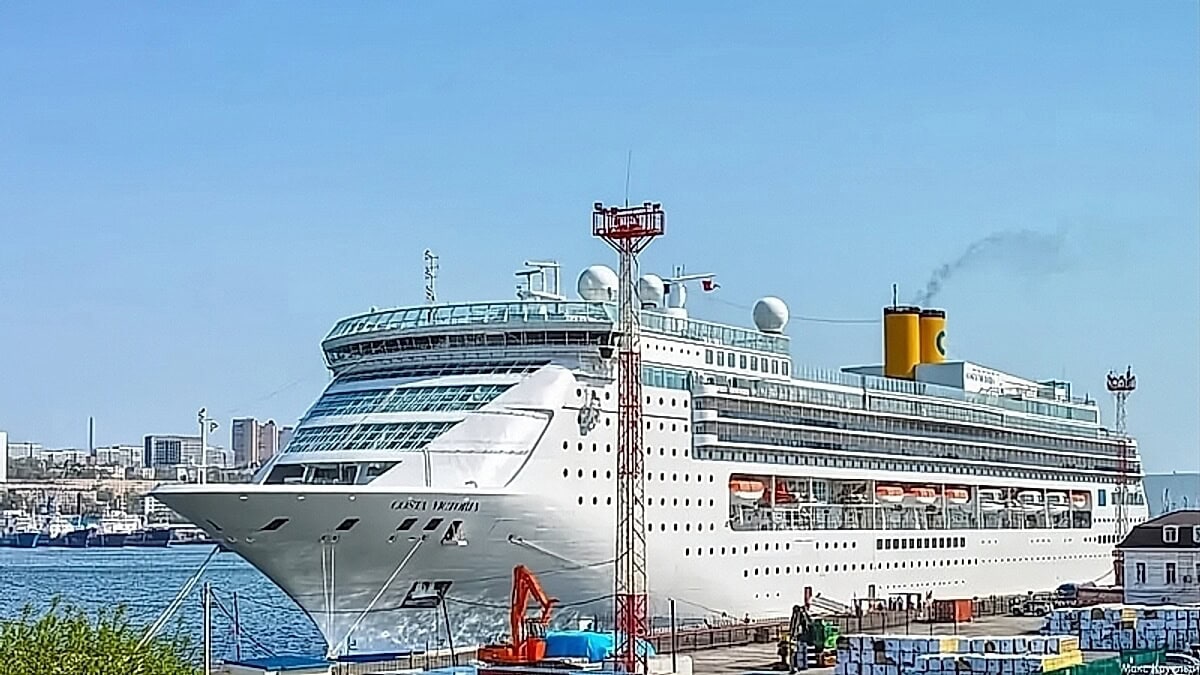 A large white cruise ship, reminiscent of the ones Carnival ships sold, is docked in a port with buildings in the background under a clear blue sky.