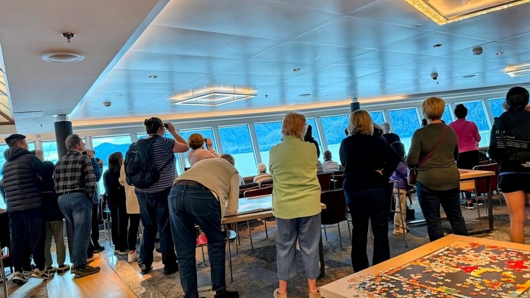 People in a room with large windows overlooking a scenic view, many standing and taking photos. A table with a puzzle in progress is in the foreground. It feels like we're cruising through the Inside Passage of Alaska, surrounded by natural beauty at every turn.