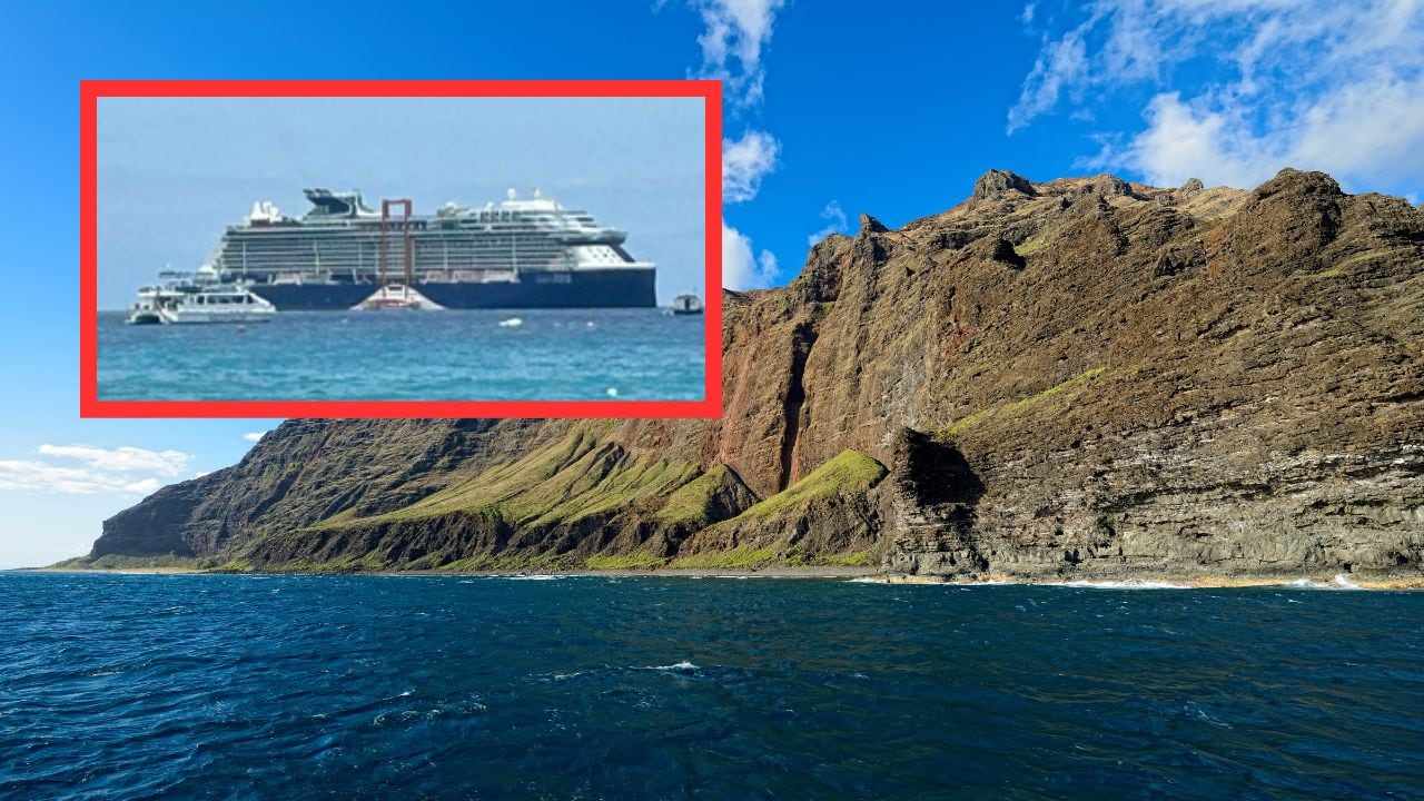 A large cruise ship is anchored off the shore near a rocky, mountainous coastline under a clear blue sky with a few scattered clouds.