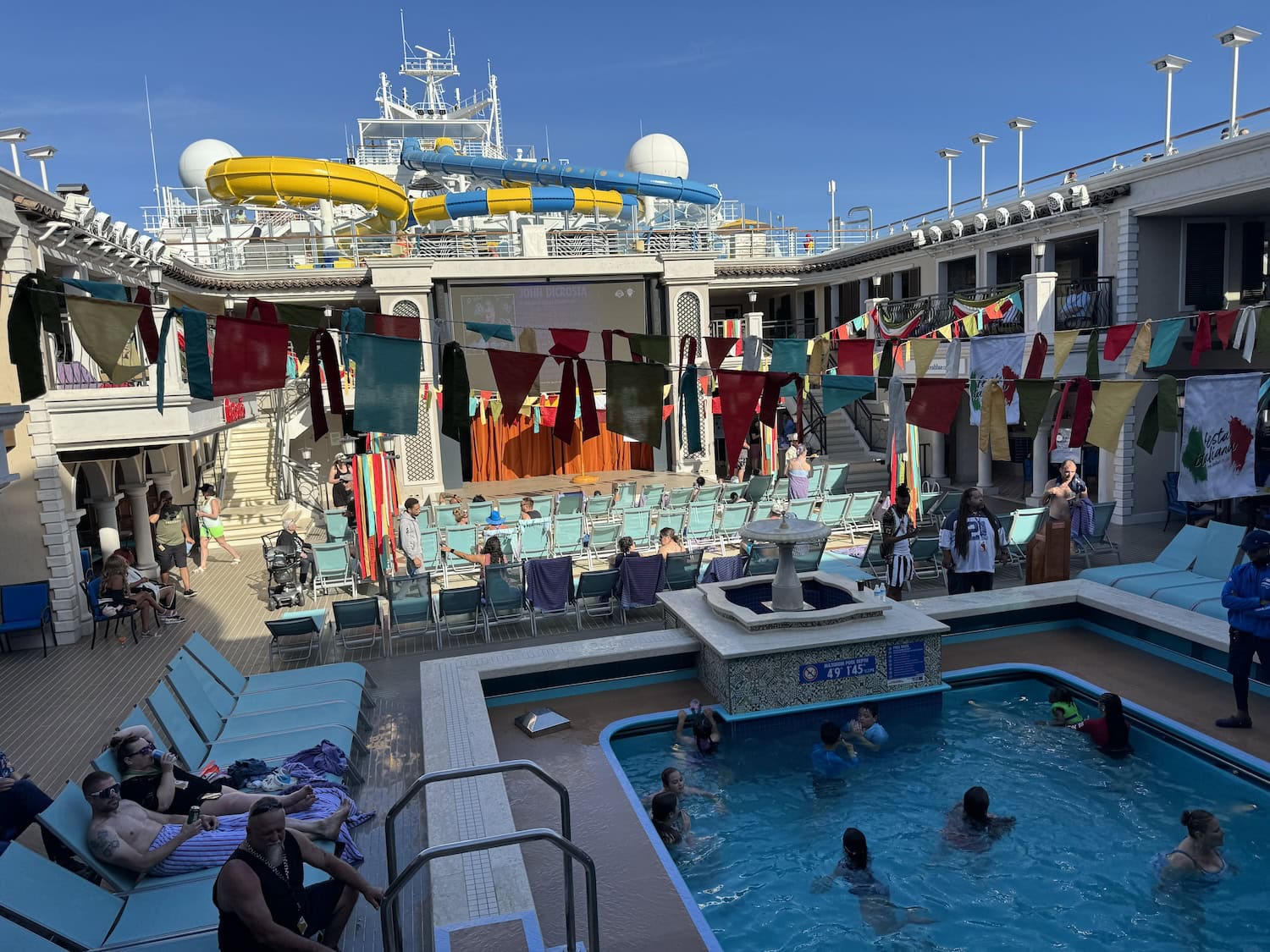 Cruise ship deck with passengers enjoying a pool, surrounded by colorful flags and deck chairs, with water slides visible in the background.