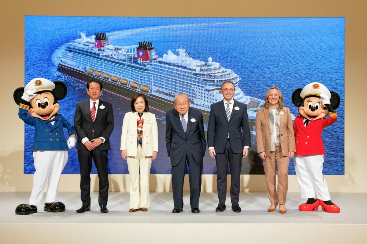 A group of six people, including two costumed Disney characters, stand in front of a backdrop featuring a cruise ship. The people are dressed formally, while the Disney characters wear maritime-themed outfits that add a touch of Tokyo charm.