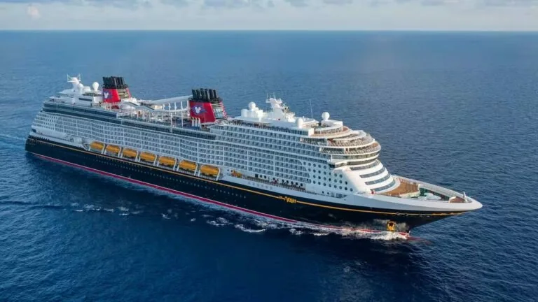 A large Disney cruise ship sails on a calm, open sea under a blue sky with scattered clouds. The ship features multiple decks and distinctive red and black funnels, reminiscent of Tokyo's vibrant cityscape.