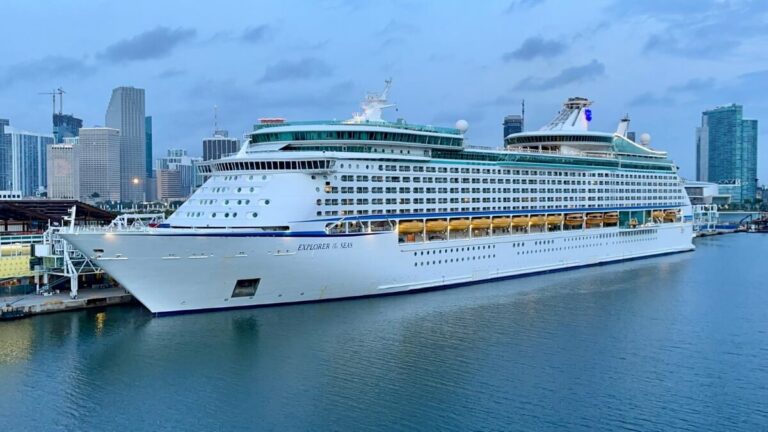 The Explorer of the Seas, a large white cruise ship, is docked at a port in a cityscape during the evening, with buildings and a cloudy sky in the background. This Mediterranean Cruise scene could easily be featured in our 2024 Podcast.