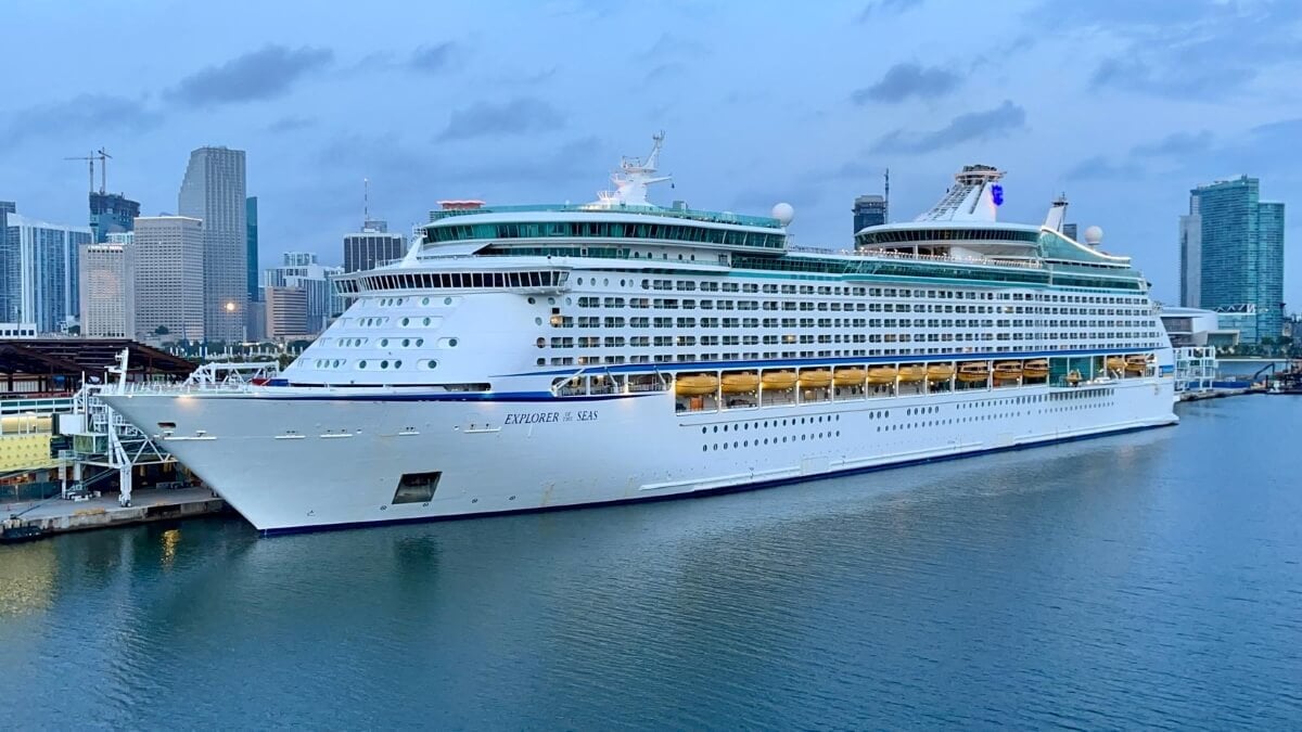 The Explorer of the Seas, a large white cruise ship, is docked at a port in a cityscape during the evening, with buildings and a cloudy sky in the background. This Mediterranean Cruise scene could easily be featured in our 2024 Podcast.