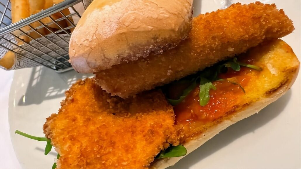 Close-up of a crispy fish sandwich with lettuce and sauce on a white plate, reminiscent of an Alaskan trip, accompanied by a small basket of golden fries.