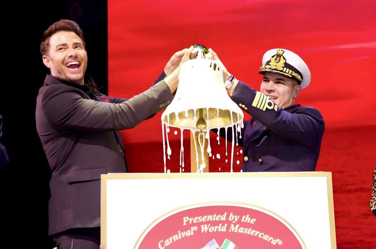 Two men on stage, one in a suit and the other in a naval officer uniform, joyfully pouring a large bottle over a ceremonial bell at a carnival event.