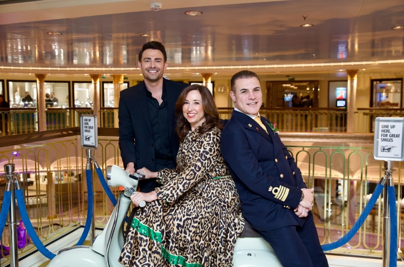 Three people, two men and one woman, smiling and sitting on a carousel horse inside a luxurious shopping mall.