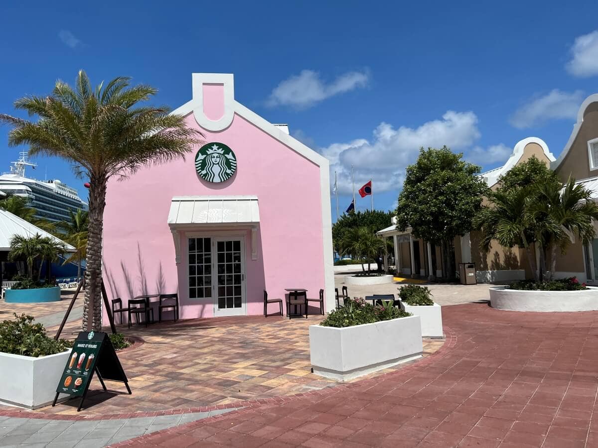 A pink building with a Starbucks logo, outdoor seating, and a cobblestone patio, situated in a sunny location with palm trees and other buildings in the background.