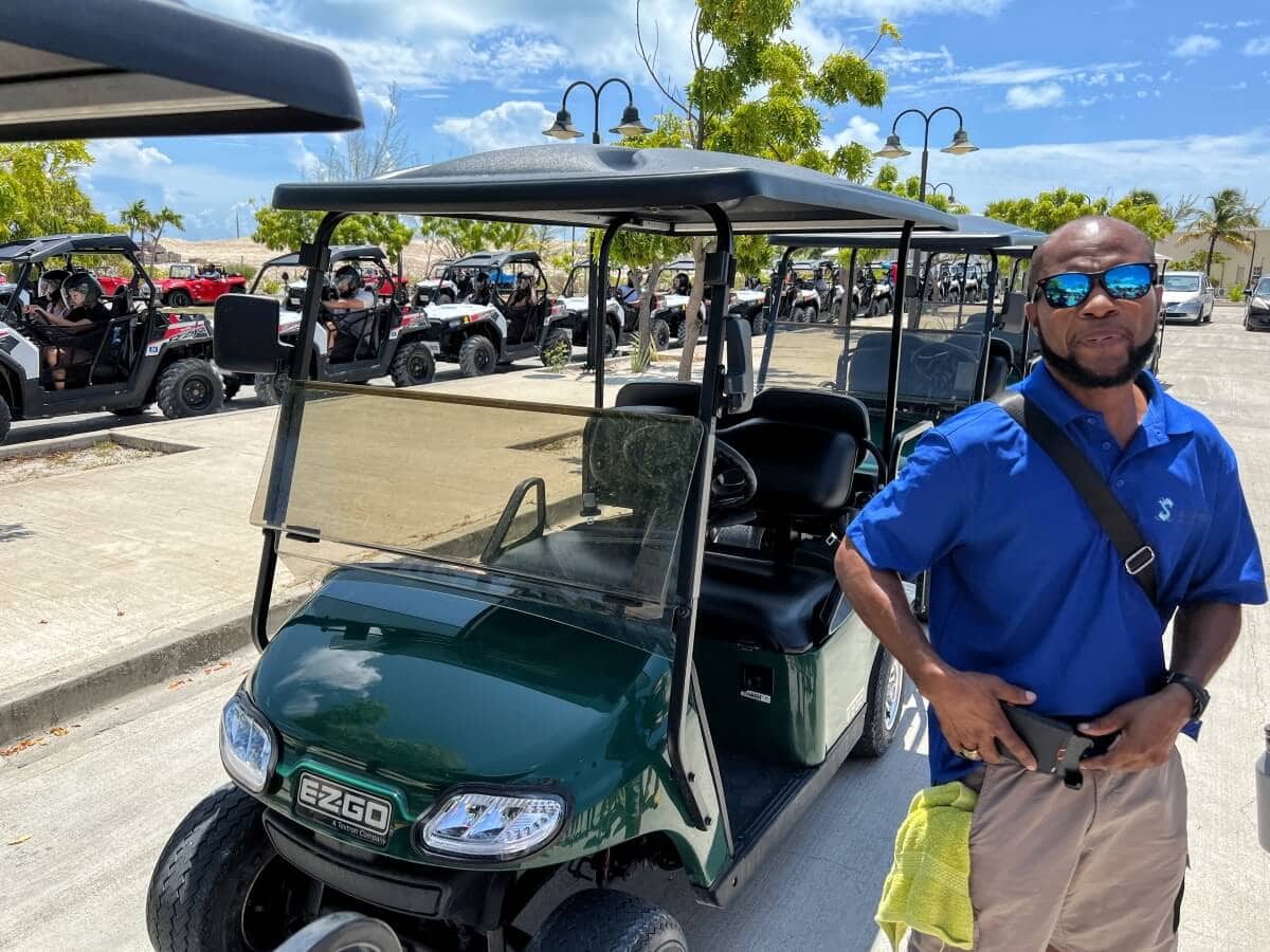 A person in a blue shirt stands near a green E-Z-GO golf cart. Several other golf carts are lined up in the background on a sunny day.