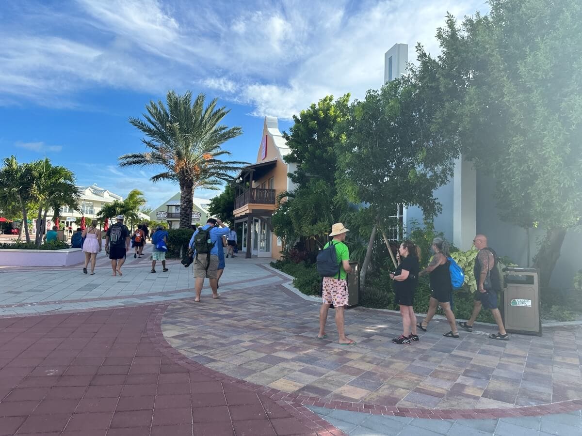 People walk and stand in a plaza with palm trees, buildings, and clear blue skies. Some carry bags and wear hats in a tropical setting.