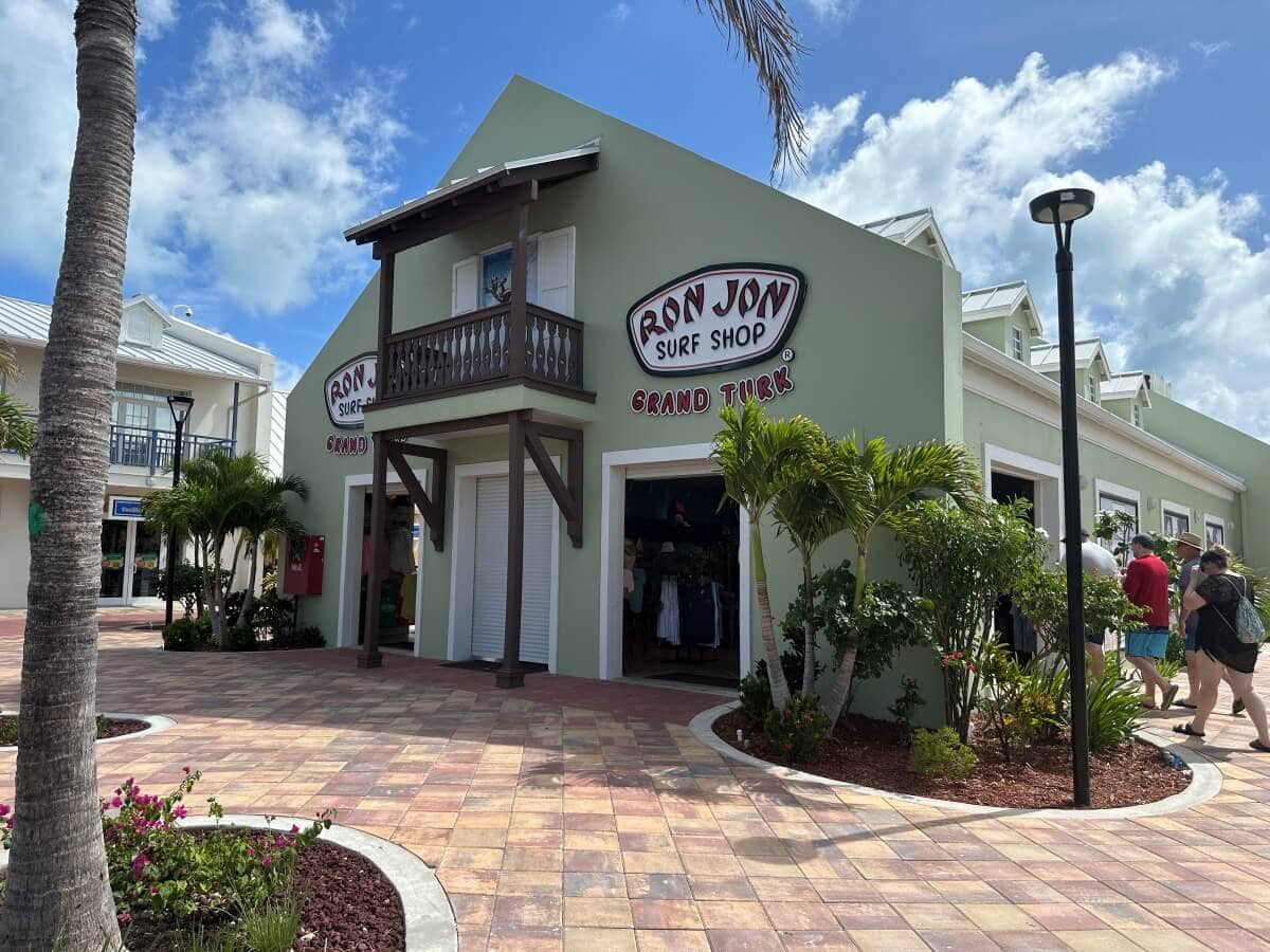 A Ron Jon Surf Shop with green walls and brown trim, located in a tropical area with palm trees and a clear sky. Two entrances are visible, with customers near the right entrance.