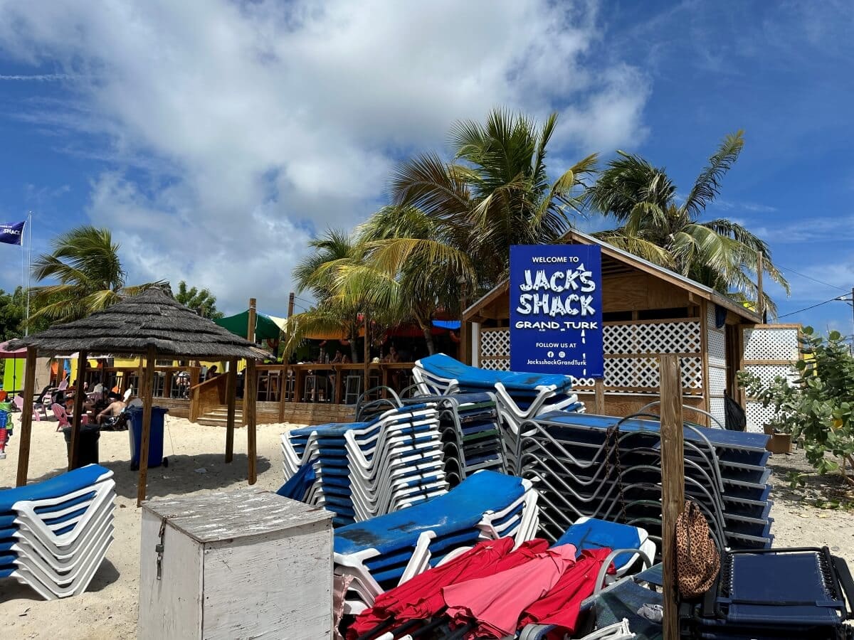 Stacks of lounge chairs and parasols are in the foreground on a beach near a wooden building with a sign that reads 