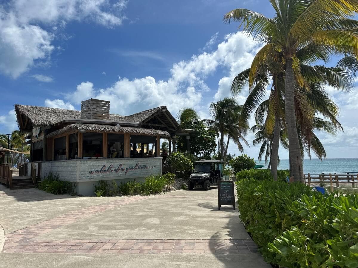 A beachside restaurant with a thatched roof and a surrounding garden path. Palm trees are nearby, and the ocean is visible in the background under a partly cloudy sky.