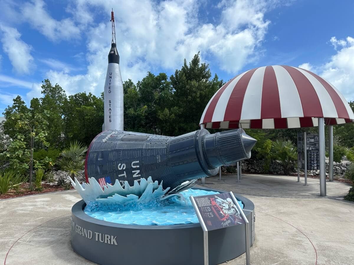 A replica of a space capsule displayed on a pool of water with a striped canopy structure nearby, and a tall rocket model in the background under a partly cloudy sky.