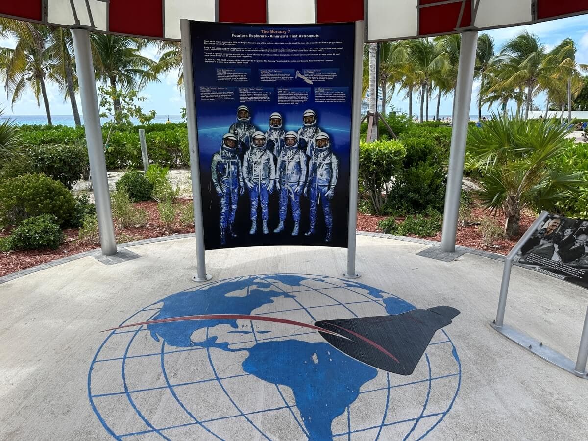 A display under a pavilion shows six figures in astronaut suits and accompanying text, with a graphic of Earth and a spacecraft projected on the ground below. Trees and the ocean are in the background.