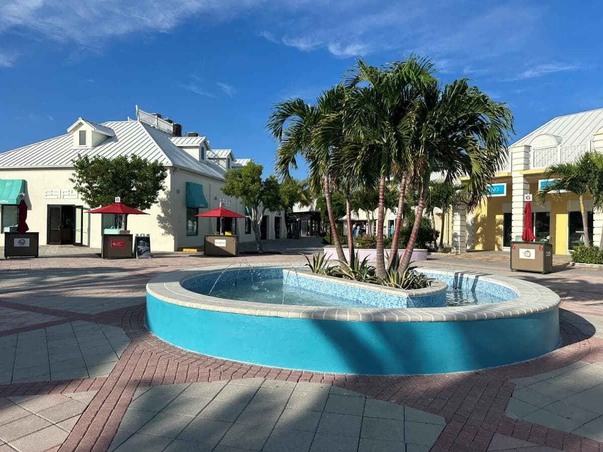 A sunny outdoor plaza features a round blue fountain surrounded by palm trees. Several shops with white facades and red umbrellas are visible in the background.