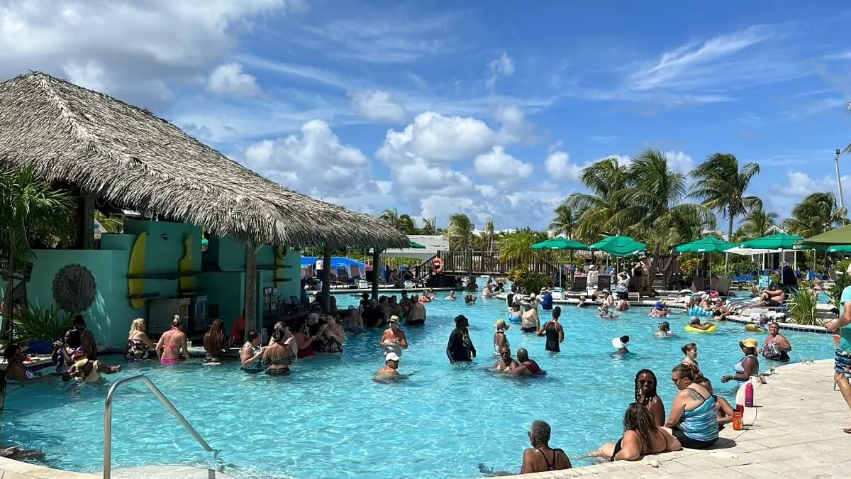 People are enjoying a sunny day in a large outdoor pool with a thatched-roof bar, surrounded by green umbrellas and palm trees.