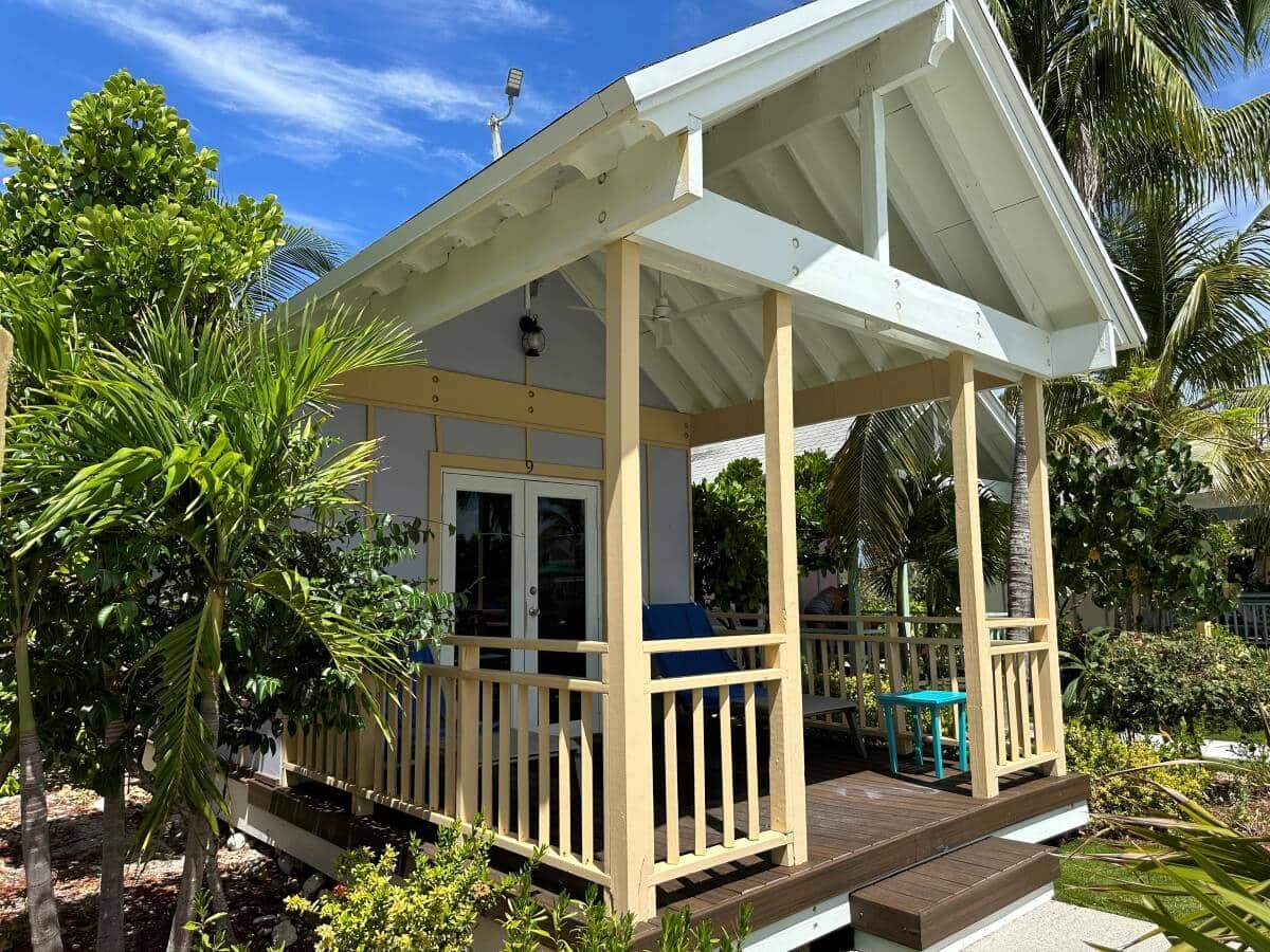 A small, white bungalow with a covered porch stands amidst lush greenery and tropical plants. The sky is clear and blue, with a few clouds visible.