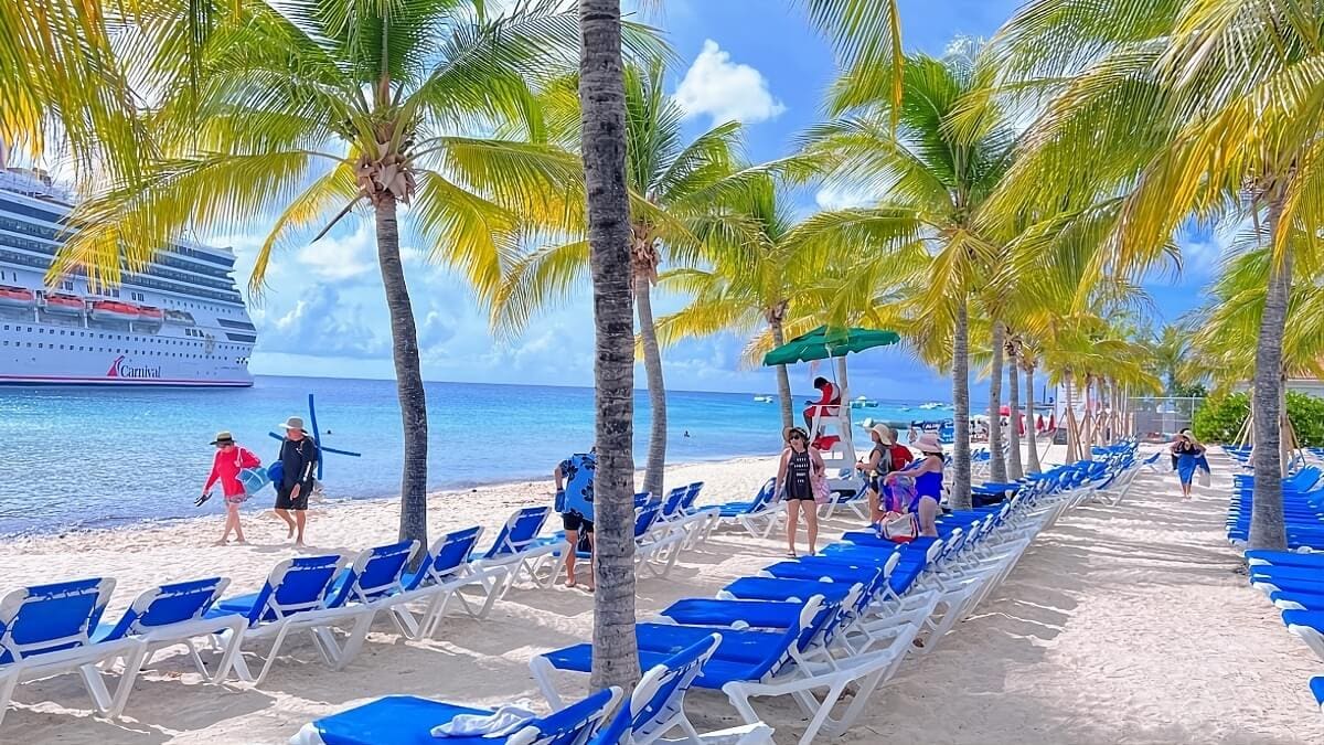 A beach with chairs and palm trees near a cruise ship.