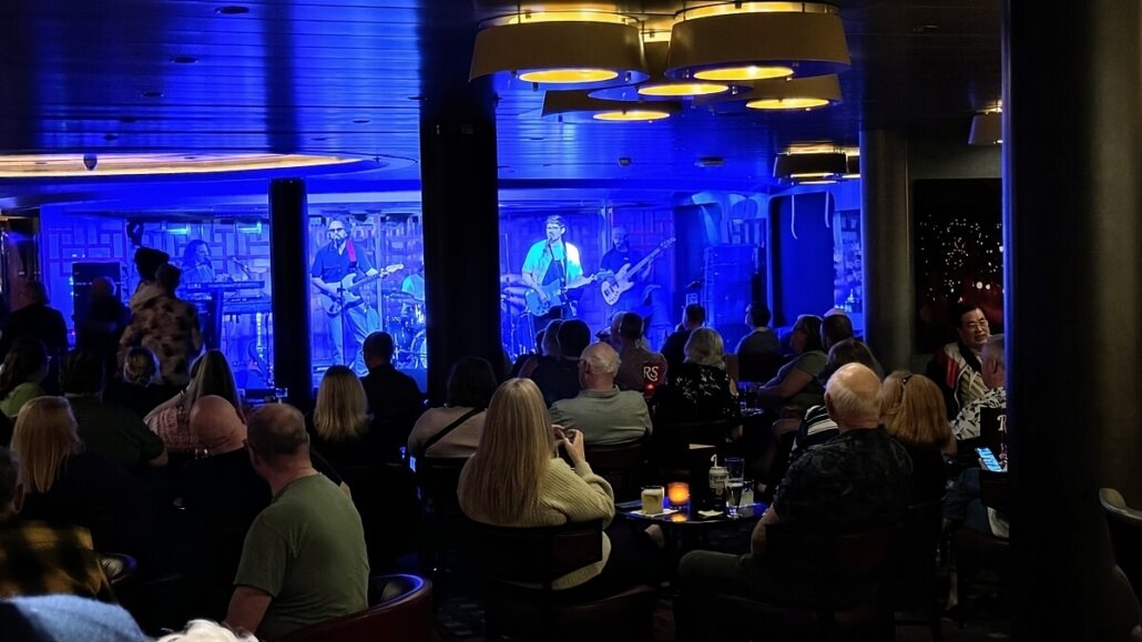 A live band performs on stage in a dimly lit venue with blue lighting, much like the intimate lounges on an Alaska trip through the Inside Passage. An audience sits at tables, watching the performance. Some people are standing, and various drinks can be seen on the tables.