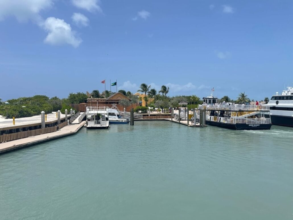 A marina with several boats docked offers a picturesque view. A building with flags on poles stands in the background, surrounded by palm trees. The sky is clear and blue, and the water is calm—just one of the many serene scenes you can enjoy while exploring Things to Do in Half Moon Cay.