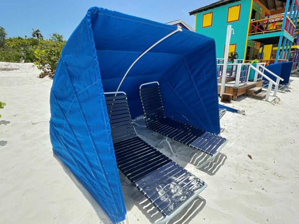 Two blue lounge chairs with a canopy shade are set up on a sandy beach in front of colorful beach huts, showcasing just one of the many relaxing things to do in Half Moon Cay.