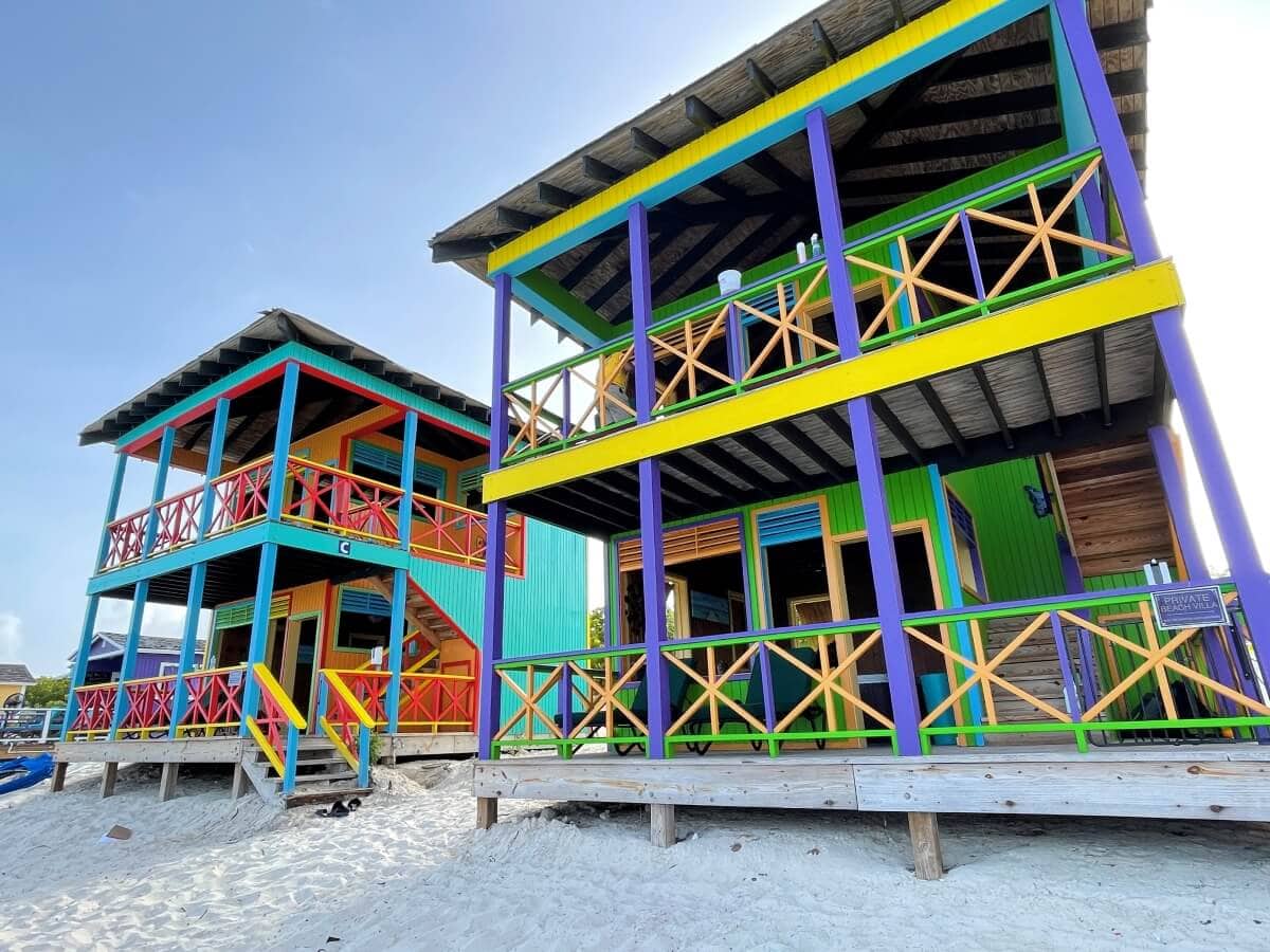 Two colorful, two-story beach houses with balconies and wooden railings stand on a sandy beach under a clear sky, just one of the many things to do in Half Moon Cay.