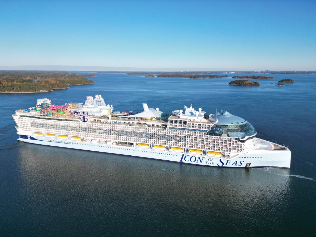 A large cruise ship named "Icon of the Seas" sails on calm water with small islands and a clear blue sky in the background. The vessel, boasting multiple decks with various amenities, raises the question: Can a cruise ship tip over?