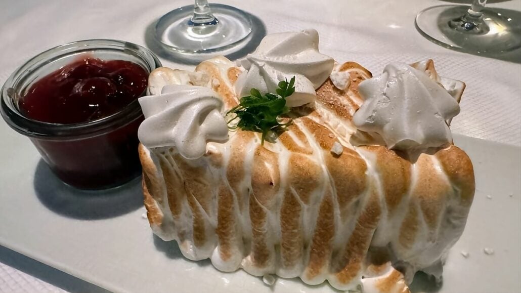 A dessert with meringue topping served on a white plate, alongside a small glass container of red cherry sauce garnished with greenery, evokes the elegance of an Alaska trip cruising through the Inside Passage.