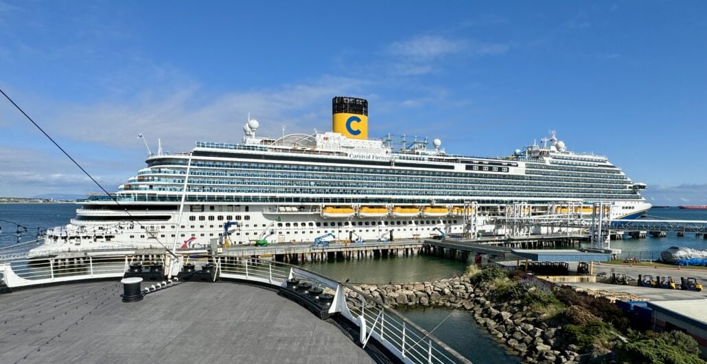 Large cruise ship docked at a pier under a clear blue sky, with visible Carnival Firenze logo on the funnel and people on the deck.