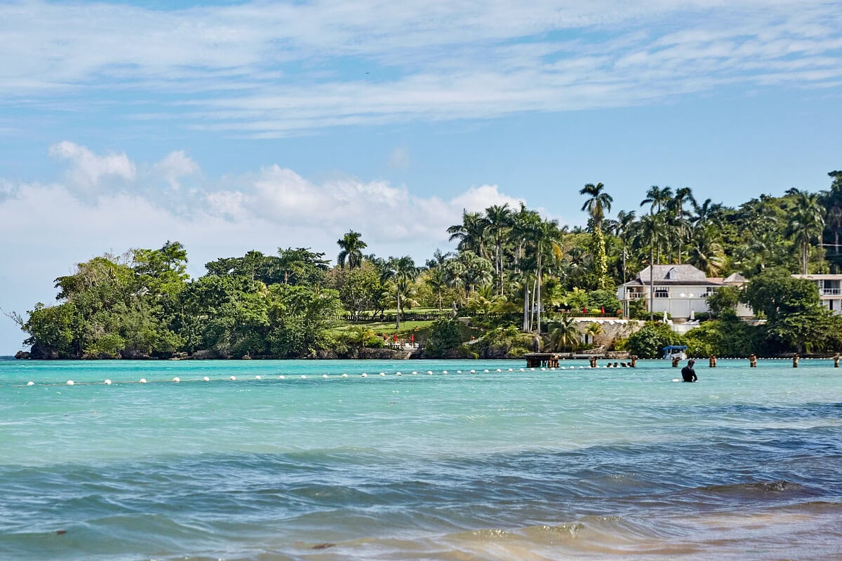 A tropical coastline with clear turquoise water, a sandy beach in the foreground, and greenery and houses on the hilly shoreline under a partly cloudy sky.